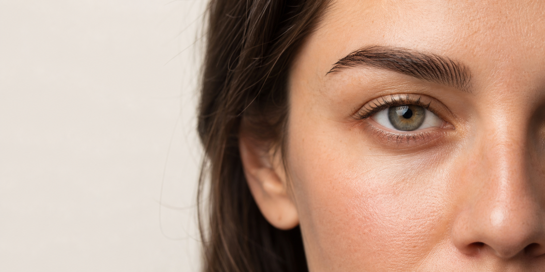 Close-up do terço superior do rosto de uma mulher, com foco na sobrancelha e na pálpebra superior, pele natural e iluminação suave lateral